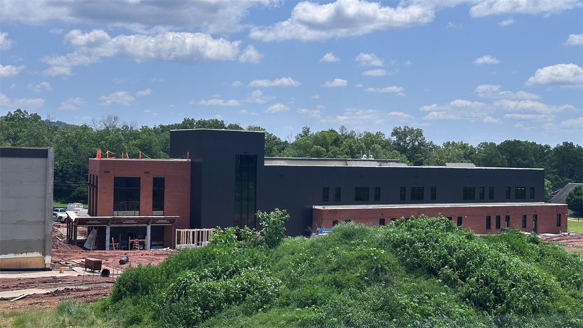 A view from the distance of large building, mostly made of brick), under construction, with green trees in the foreground.